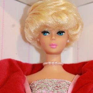 Peut inclure: Une poupée blonde portant une veste en velours rouge et une robe à paillettes rose et argent. La poupée a les yeux bleus et les lèvres roses.