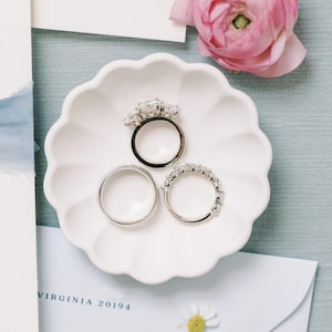 Puede incluir: Tres alianzas de boda plateadas en un platillo blanco. La banda central tiene tres diamantes grandes. El platillo está sobre una superficie azul claro.