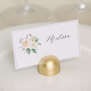 May include: A place setting with a white plate, a gold-colored place card holder, and a place card with the name "Melissa" and a floral design. Two wine glasses are in the background.