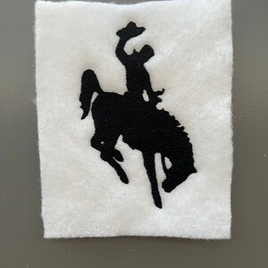 May include: Black silhouette of a cowboy riding a bucking horse, embroidered on a white felt patch.