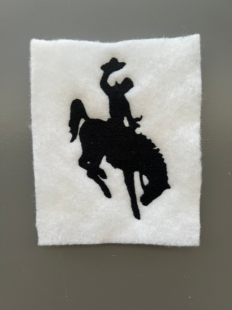May include: Black silhouette of a cowboy riding a bucking horse, embroidered on a white felt patch.