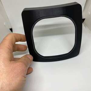 May include: A black plastic frame with a rounded top and a square opening. The frame is designed to hold a small object, such as a phone or a tablet.