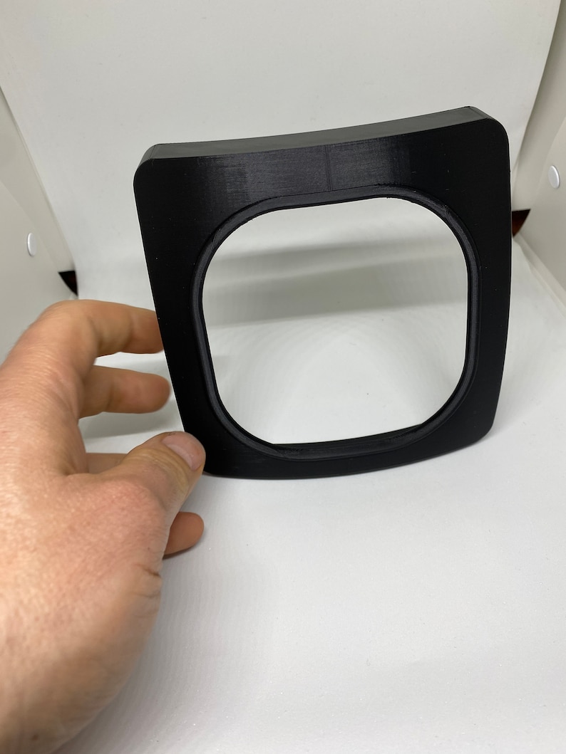 May include: A black plastic frame with a rounded top and a square opening. The frame is designed to hold a small object, such as a phone or a tablet.