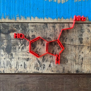 Può includere: Un modello 3D rosso della struttura chimica della serotonina, un neurotrasmettitore, su una superficie di legno. Il modello è in plastica e presenta un design dettagliato. Lo sfondo è una superficie di legno dipinta di blu.