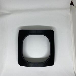 May include: A black plastic ring with a circular opening in the center. The ring is designed to fit around a camera lens.