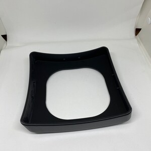 May include: Black plastic base with a circular cutout in the center. The base has a slightly curved shape and is designed to hold a round object.