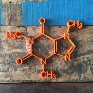 Puede incluir: Un modelo 3D naranja de una molécula de cafeína sobre un fondo de madera. La molécula está hecha de plástico y tiene la fórmula química C8H10N4O2.