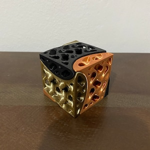 Può includere: Un cubo stampato in 3D con finitura nera, dorata e rame. Il cubo presenta un design geometrico a forma di reticolo.