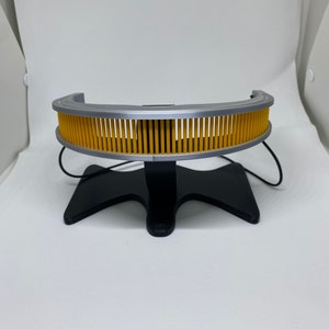 May include: A silver and yellow headband with a stand. The headband has a series of yellow vertical bars.