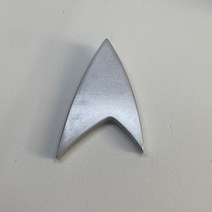 May include: A silver-coloured Starfleet insignia, a triangular shape with a point at the top and a curved bottom, is shown against a white background.