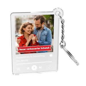 Könnte beinhalten: Ein weißer Acryl-Schlüsselanhänger mit einem Foto eines Paares vor einer Stadtkulisse. Das Foto ist mit einem roten Banner mit der Aufschrift "Neuer verbesserter Schutz!" und einer weißen Musikplayer-Oberfläche mit dem Text "Happiness is travelling the world" gerahmt.