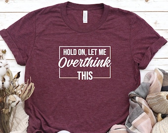 May include: Heather maroon t-shirt with the text "HOLD ON, LET ME Overthink THIS" in a white rectangular box. The shirt is paired with white sneakers, a cream-colored sweater, and a scarf. The words "HEATHER MAROON" are in the top left corner.