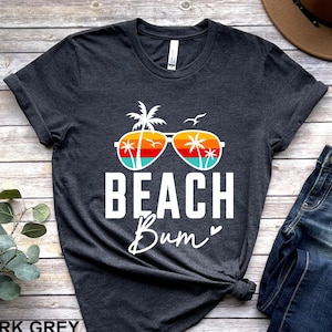 Pode incluir: Camiseta cinza escuro com efeito de mescla com um gr&aacute;fico branco de &oacute;culos de sol com palmeiras dentro das lentes. O texto "Beach Bum" est&aacute; abaixo dos &oacute;culos de sol.