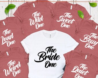 May include: Set of seven t-shirts with different phrases printed on them. The shirts are in shades of maroon and white. The phrases include "The Wild One", "The Sweet One", "The Loud One", "The Bossy One", "The Weird One", "The Quiet One", and "The Bride One".