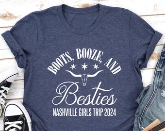 May include: A navy blue t-shirt with white text that reads "Boots, Booze, and Besties Nashville Girls Trip 2024". The text is in a western-style font with a skull and crossbones graphic.
