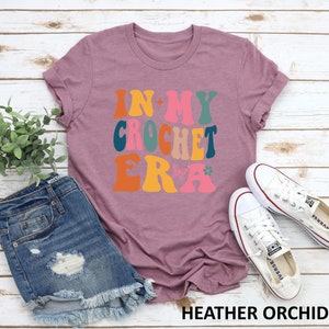 May include: A heather orchid colored t-shirt with the text "In My Crochet Era" in a retro font with colorful letters and a butterfly graphic.