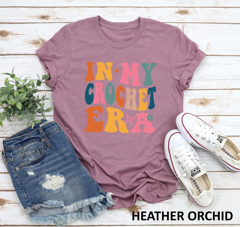 May include: A heather orchid colored t-shirt with the text "In My Crochet Era" in a retro font with colorful letters and a butterfly graphic.
