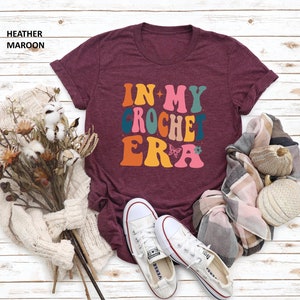 May include: A maroon t-shirt with the text "In My Crochet Era" in a retro font with colorful letters and a butterfly graphic.