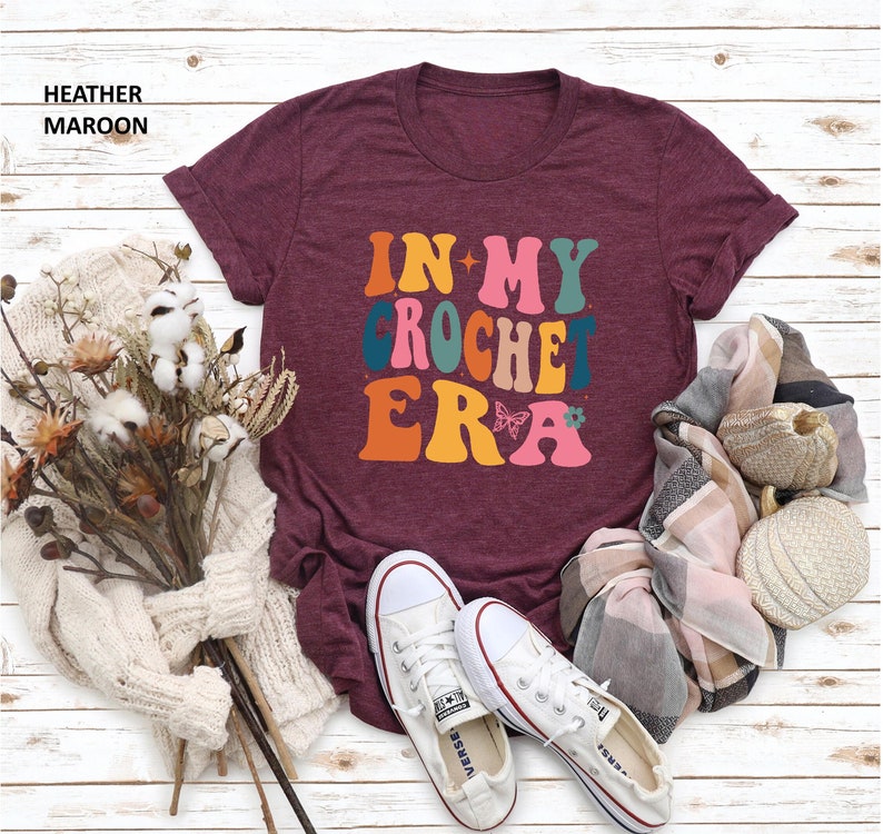 May include: A maroon t-shirt with the text "In My Crochet Era" in a retro font with colorful letters and a butterfly graphic.