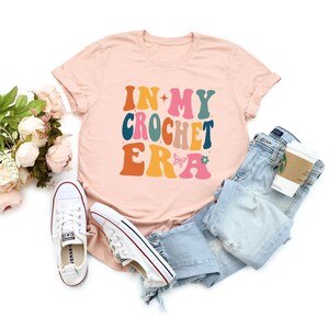 May include: A pink t-shirt with the text "In My Crochet Era" in colorful retro-style lettering. The shirt is paired with blue jeans, white sneakers, and a coffee cup.