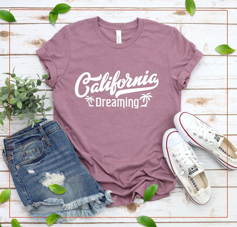 May include: A mauve t-shirt with the text "California Dreaming" in white with palm tree graphics. The shirt is laid out on a white wood surface with a pair of white canvas shoes and denim shorts.