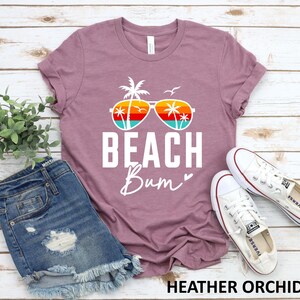 Pode incluir: Uma camiseta cor orqu&iacute;dea com um gr&aacute;fico branco de um &oacute;culos de sol com palmeiras refletidas nas lentes. O texto "Beach Bum" est&aacute; impresso em branco abaixo dos &oacute;culos de sol.