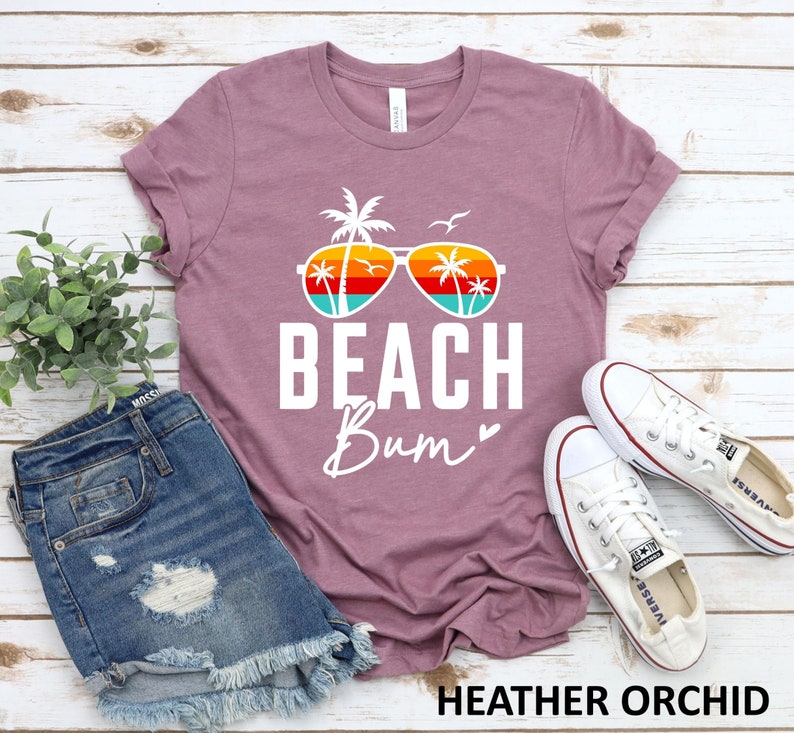 Pode incluir: Uma camiseta cor orqu&iacute;dea com um gr&aacute;fico branco de um &oacute;culos de sol com palmeiras refletidas nas lentes. O texto "Beach Bum" est&aacute; impresso em branco abaixo dos &oacute;culos de sol.