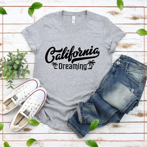 May include: A gray t-shirt with black text that reads "California Dreaming" with palm trees on either side of the text. The shirt is laid out on a wooden surface with a pair of white sneakers and blue jeans.