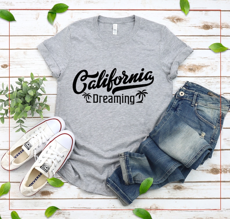 May include: A gray t-shirt with black text that reads "California Dreaming" with palm trees on either side of the text. The shirt is laid out on a wooden surface with a pair of white sneakers and blue jeans.