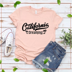 May include: A pink t-shirt with black text that reads "California Dreaming" with palm trees on either side of the text. The t-shirt is laid out on a white wooden surface with a pair of white sneakers and denim shorts.