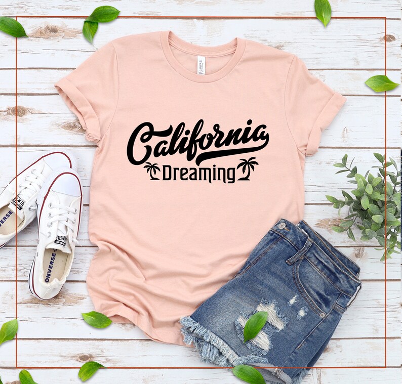 May include: A pink t-shirt with black text that reads "California Dreaming" with palm trees on either side of the text. The t-shirt is laid out on a white wooden surface with a pair of white sneakers and denim shorts.