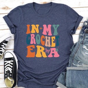 May include: A navy blue t-shirt with the text "In My Crochet Era" in colorful retro-style lettering. The text is decorated with a butterfly and a flower.