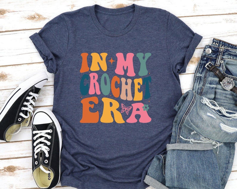 May include: A navy blue t-shirt with the text "In My Crochet Era" in colorful retro-style lettering. The text is decorated with a butterfly and a flower.
