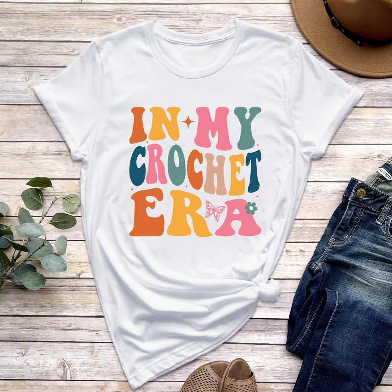 May include: White t-shirt with the text "In My Crochet Era" in colorful, retro-style lettering. A butterfly and flower are included in the design.
