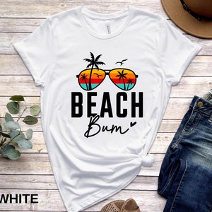 Pode incluir: Camiseta branca com um gr&aacute;fico de &oacute;culos de sol com palmeiras e um p&ocirc;r do sol. O texto "Beach Bum" &eacute; impresso abaixo dos &oacute;culos de sol.