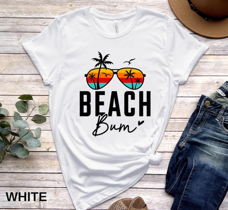 Pode incluir: Camiseta branca com um gr&aacute;fico de &oacute;culos de sol com palmeiras e um p&ocirc;r do sol. O texto "Beach Bum" &eacute; impresso abaixo dos &oacute;culos de sol.
