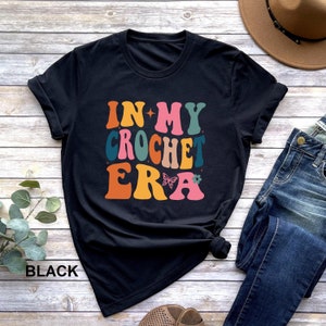 May include: Black t-shirt with the text "In My Crochet Era" in colorful retro-style lettering. The text is decorated with a butterfly and a flower.