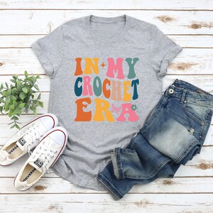 May include: A gray t-shirt with the text "In My Crochet Era" in colorful, retro-style lettering. The shirt is paired with blue jeans and white sneakers.