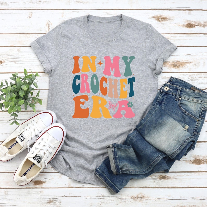May include: A gray t-shirt with the text "In My Crochet Era" in colorful, retro-style lettering. The shirt is paired with blue jeans and white sneakers.