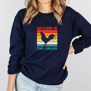 May include: A navy blue sweatshirt with a retro-style graphic of a rooster and the text "I'm fluent in fowl language".