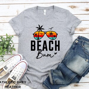Pode incluir: Uma camiseta cinza com um gr&aacute;fico de &oacute;culos de sol com palmeiras refletidas nas lentes. O texto "Beach Bum" est&aacute; impresso abaixo dos &oacute;culos de sol.