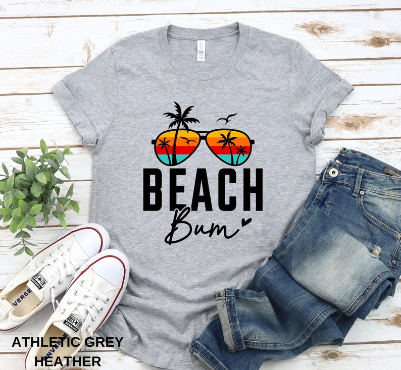 Pode incluir: Uma camiseta cinza com um gr&aacute;fico de &oacute;culos de sol com palmeiras refletidas nas lentes. O texto "Beach Bum" est&aacute; impresso abaixo dos &oacute;culos de sol.