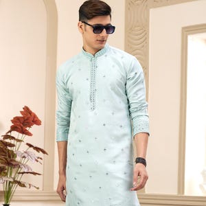 May include: A light blue long-sleeved kurta with a white embroidered design. The kurta has a mandarin collar and a button closure. The model is wearing white pants and brown shoes.