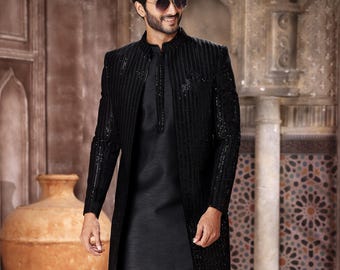 May include: A man in a black formal outfit, including a long jacket with sequin detailing, a mandarin-collared shirt, and matching pants. The ensemble is completed with black shoes with gold accents. The background features an ornate, textured wall and a large vase.