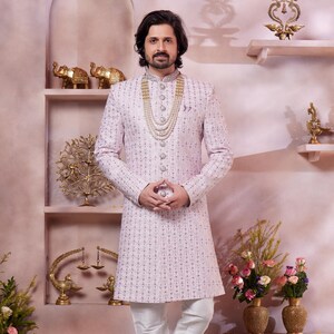 May include: A man wearing a light purple patterned kurta with a white collar and white pants. He is wearing a pearl necklace and has a white turban on his head. The background is a pink wall with gold accents.