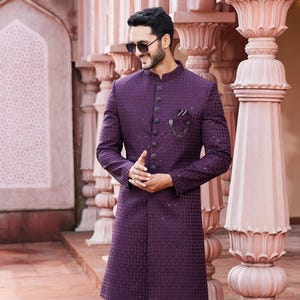 Może przedstawiać: Formalna fioletowa kurtka z teksturowanym wzorem, połączona z białymi spodniami i butami. Kurtka posiada kołnierz mandaryński i ozdobne guziki. Poszetka i łańcuszek dodają szczegółów. Strój jest odpowiedni na specjalne okazje.