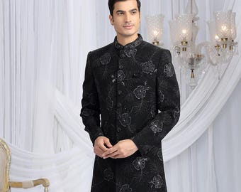 May include: A black formal suit featuring a long, floral-patterned jacket and matching trousers. The jacket has a mandarin collar and button closure. The suit is worn with black dress shoes. The background includes a decorative chair and a chandelier.
