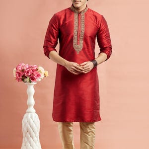 May include: A red kurta with gold embroidery on the collar and a matching pair of beige pants. The traditional Indian outfit is worn with white shoes. A decorative white vase with pink flowers is in the background.