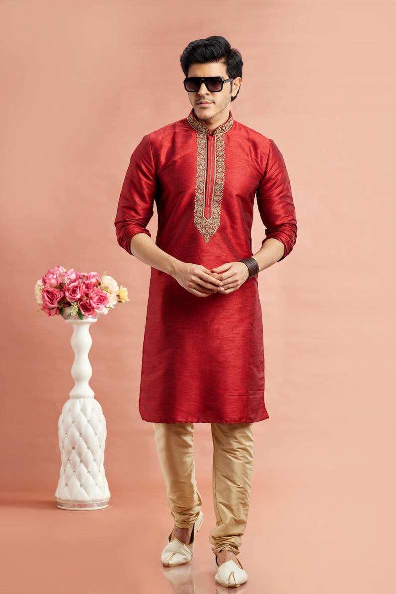 May include: A red kurta with gold embroidery on the collar and a matching pair of beige pants. The traditional Indian outfit is worn with white shoes. A decorative white vase with pink flowers is in the background.