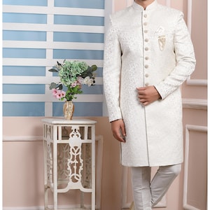Puede incluir: Una kurta blanca de manga larga con cuello mandar&iacute;n y cierre de botones. La kurta tiene un sutil estampado floral y se combina con pantalones blancos. El hombre lleva zapatos blancos y gafas.